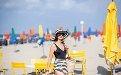 Eine stilvoll gekleidete Frau spaziert entspannt über einen sonnigen Sandstrand, umgeben von farbenfrohen Sonnenschirmen und gelben Strandstühlen.