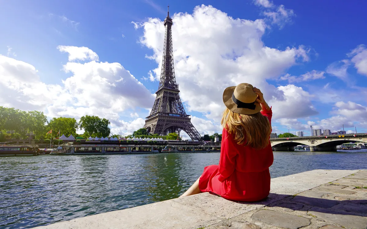 Reisende in rotem Kleid und Hut sitzt auf dem Kai der Seine und blickt auf den Eiffelturm