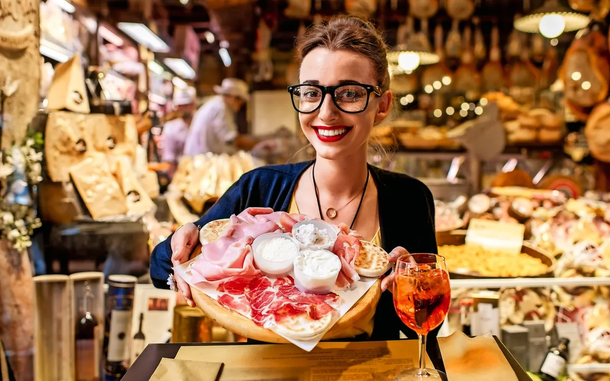 Junge Frau, die nahe dem Lebensmittelladen mit traditionellem italienischem Aperitif auf dem unscharfen Lebensmittelschaukastenhintergrund in der Bolognastadt sitzt. Bologna ist das gastronomische Zentrum Italiens