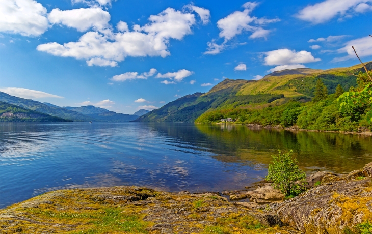 Blick auf einen See mit klarer, ruhiger Wasseroberfläche, umgeben von grünen Hügeln und Bergen unter einem blauen Himmel mit weißen Wolken. Im Vordergrund sind Felsen und etwas Vegetation zu sehen.
