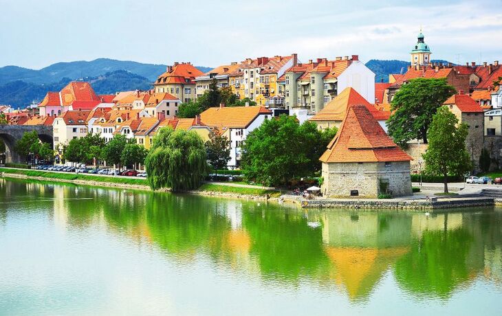 Stadtansicht mit Fluss, roten Dächern und Hügeln im Hintergrund.