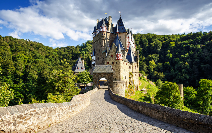 Burg Eltz mit Brücke, umgeben von Wald und blauem Himmel.