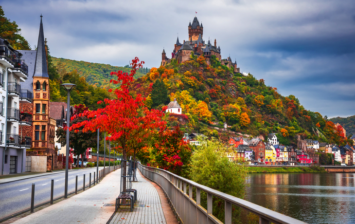 Burg auf Hügel mit Herbstlaub, Fluss und bunten Häusern im Vordergrund.