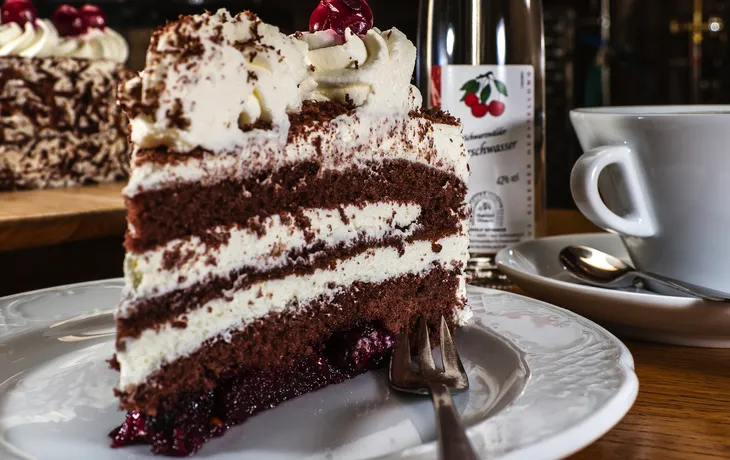 Stück Schwarzwälder Kirschtorte auf Teller mit Kaffeetasse im Hintergrund.