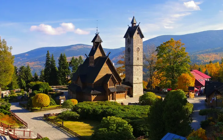 Eine alte Holzkirche und ein Steinturm in einer herbstlichen Berglandschaft.