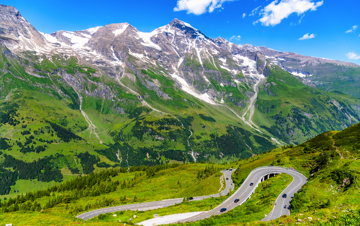 Serpentinenstraße vor einer Berglandschaft mit Schnee und grünem Tal.