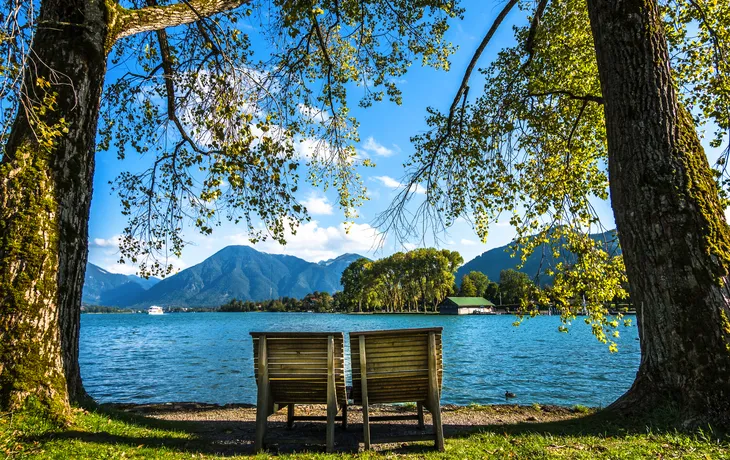 Zwei Holzbänke am Seeufer mit Blick auf Berge und Bäume.