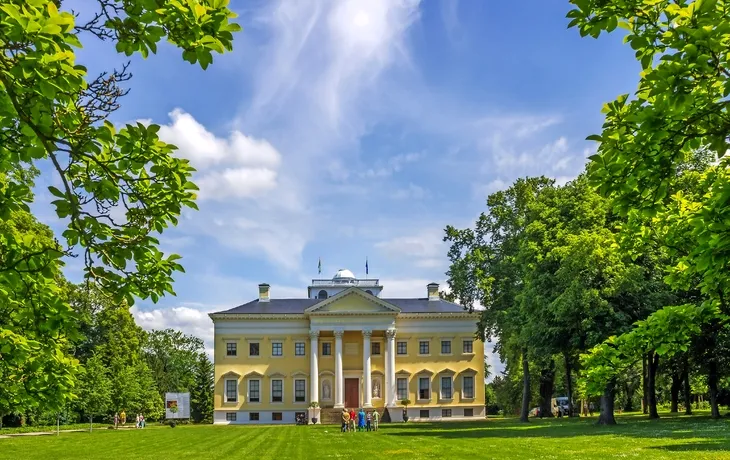 Großes gelbes Gebäude mit Säulen, umgeben von Bäumen an einem sonnigen Tag.