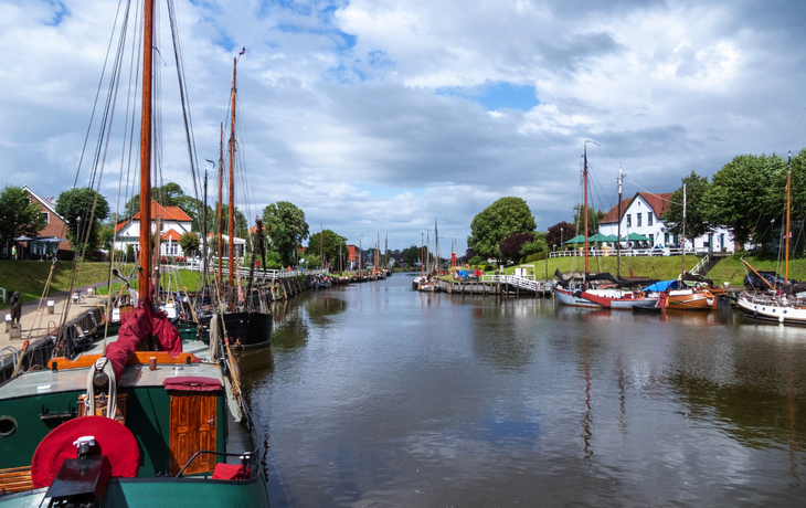 Segelboote an einem Fluss mit Uferpromenade und Häusern im Hintergrund unter bewölktem Himmel