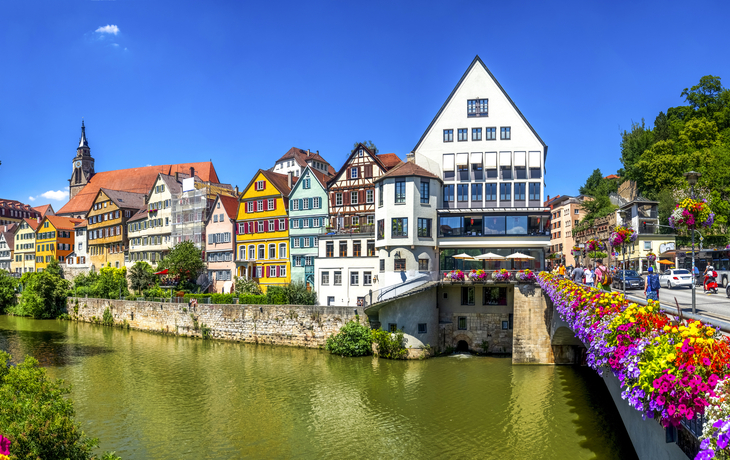Bunte Fachwerkhäuser an einem Fluss mit Blumen auf einer Brücke und blauem Himmel.