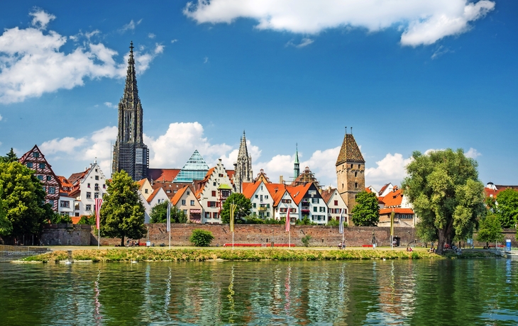 Historische Altstadt mit Fachwerkhäusern und Kirchturm am Flussufer unter blauem Himmel.