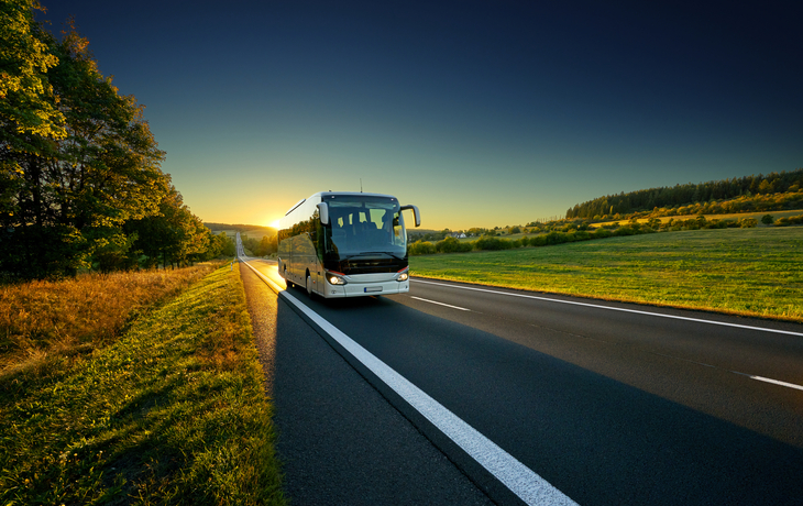 Reisebus fährt bei Sonnenuntergang auf einer Landstraße durch ländliche Umgebung.