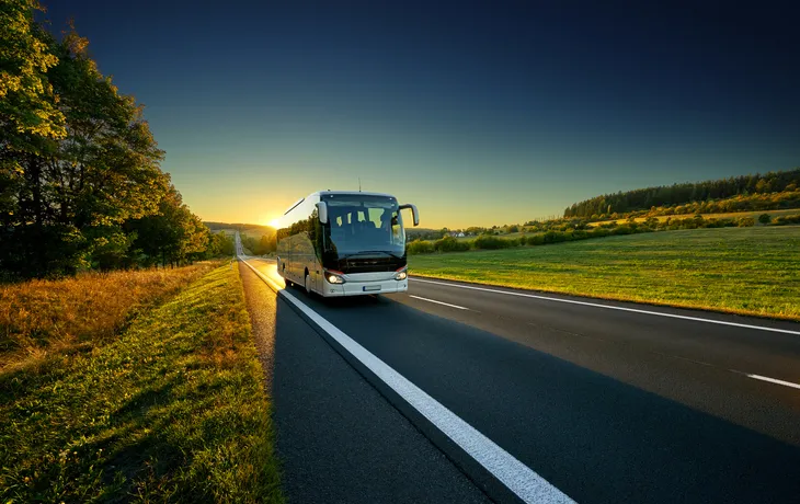 Reisebus fährt bei Sonnenuntergang auf einer Landstraße durch ländliche Umgebung.
