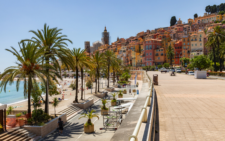Uferpromenade mit Palmen vor bunten Häusern und einem Strand bei sonnigem Wetter.