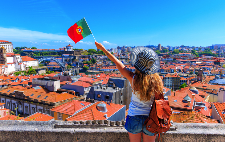 Person mit Hut schwenkt portugiesische Flagge über Stadt mit roten Dächern.